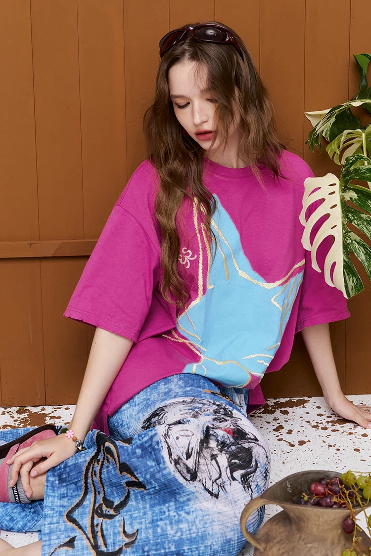 A woman wearing a bright pink oversized T-shirt with a large light blue graphic panel and gold line details, paired with a blue patterned skirt, posed in front of a brown wood-paneled wall.