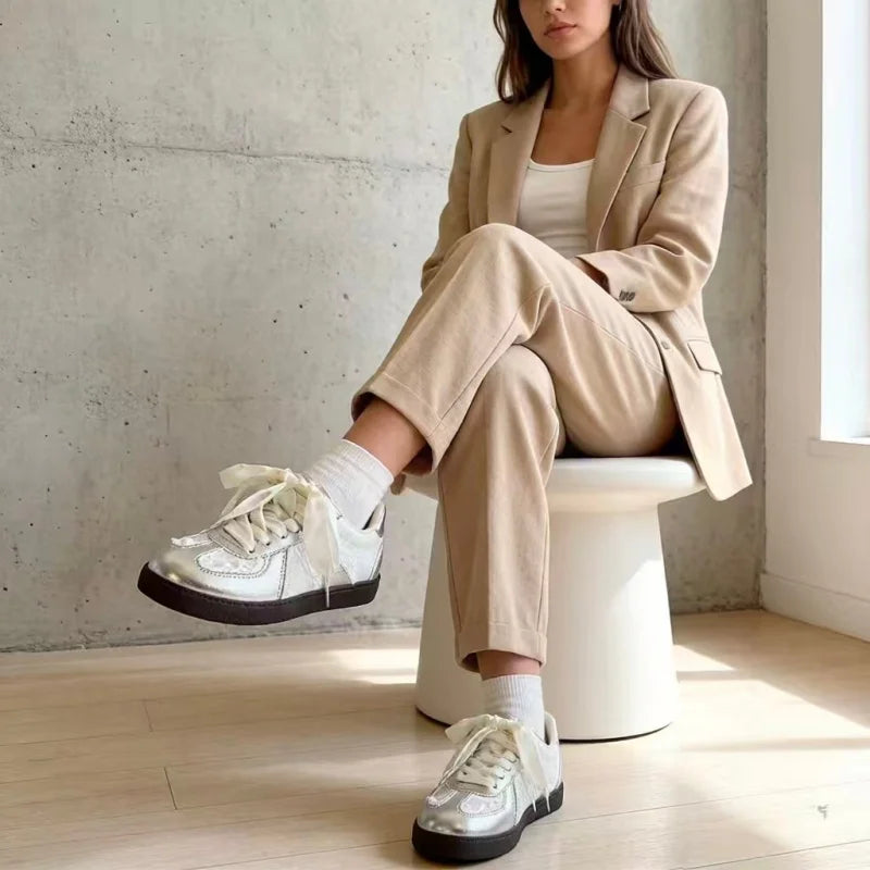 A woman sits on a white stool in a minimalist room with a concrete wall and light wood floor, wearing a beige blazer and matching trousers, white socks, and shiny silver low-top sneakers with white laces.