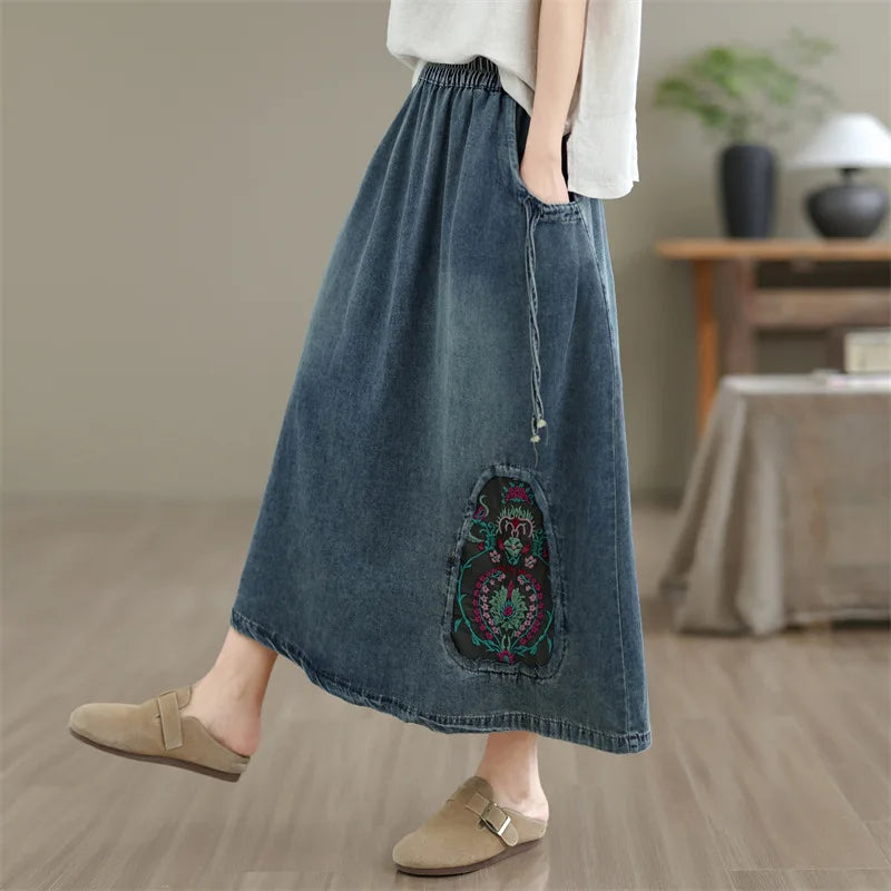A person wearing a long blue denim maxi skirt with a colorful embroidered patch detail and side pocket, paired with a white top and beige slip-on shoes in a softly lit indoor room.