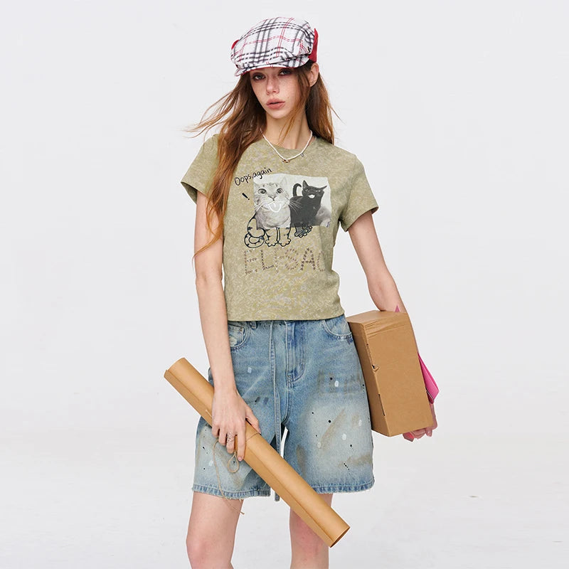 A woman modeling a short-sleeve graphic t-shirt in olive green with a cat illustration and the text 'DREAMER ELFA', paired with distressed denim shorts, a plaid cap, and holding cardboard boxes against a plain white studio background.