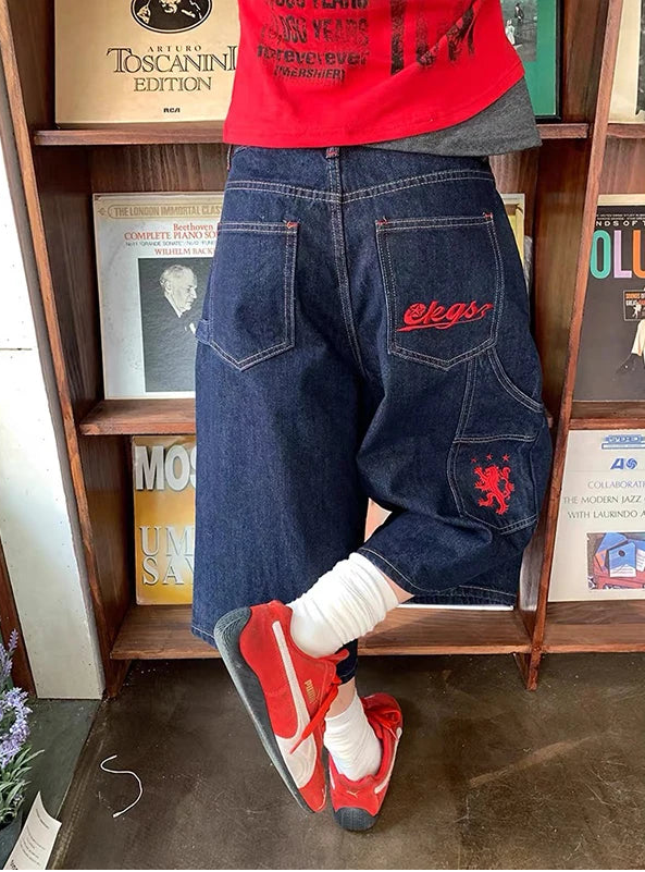A person shown from the back wearing dark blue denim shorts with red embroidered logos, a red graphic t-shirt, white socks, and red sneakers, standing in front of a wooden bookshelf.