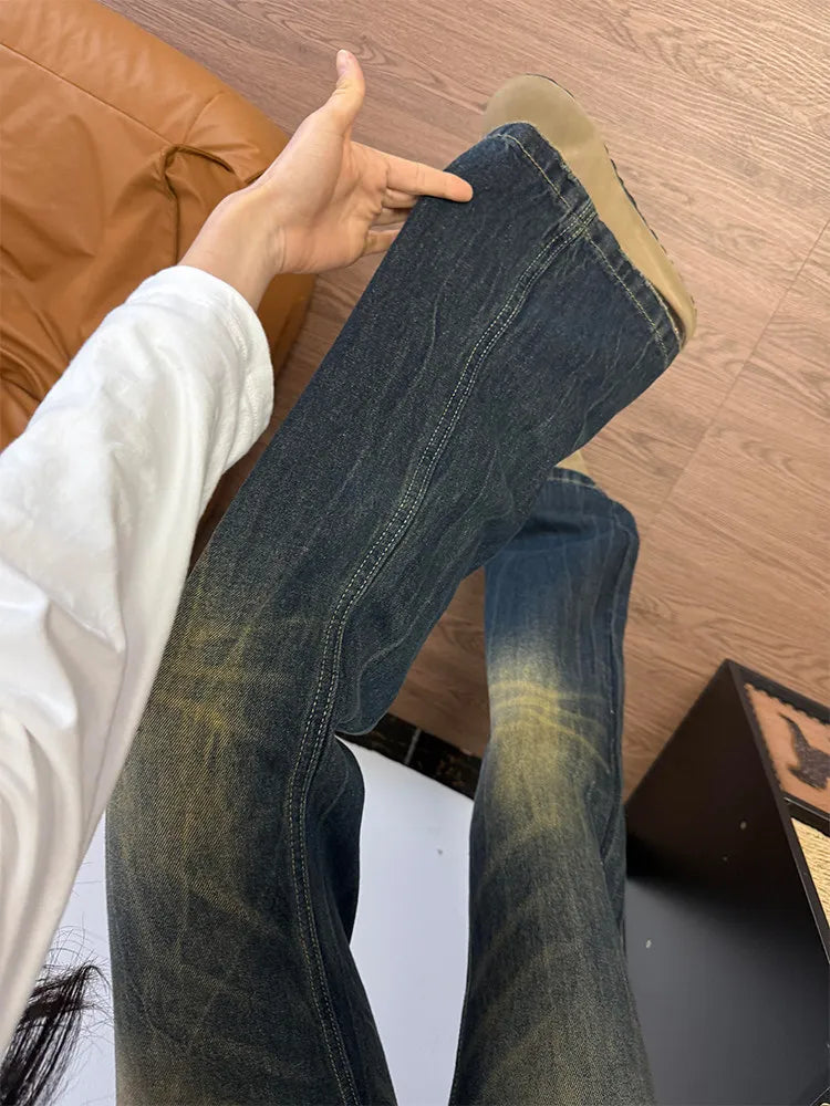 A person wearing dark blue flare jeans with a faded yellow wash, holding up one leg to show the fabric and fit. The background shows a brown leather chair and a wood floor.