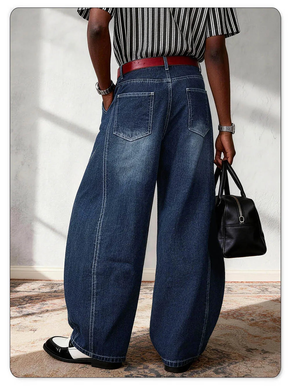 A person shown from the back wearing very wide-leg dark blue denim jeans with a red belt, a black-and-white striped shirt, and black-and-white loafers, while holding a black handbag against a light wall.
