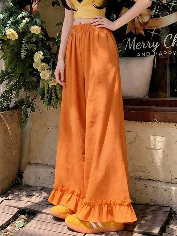 A person wearing bright orange wide-leg ruffled pants with a matching yellow crop top and yellow shoes, standing outdoors near a rustic wall and potted flowers.
