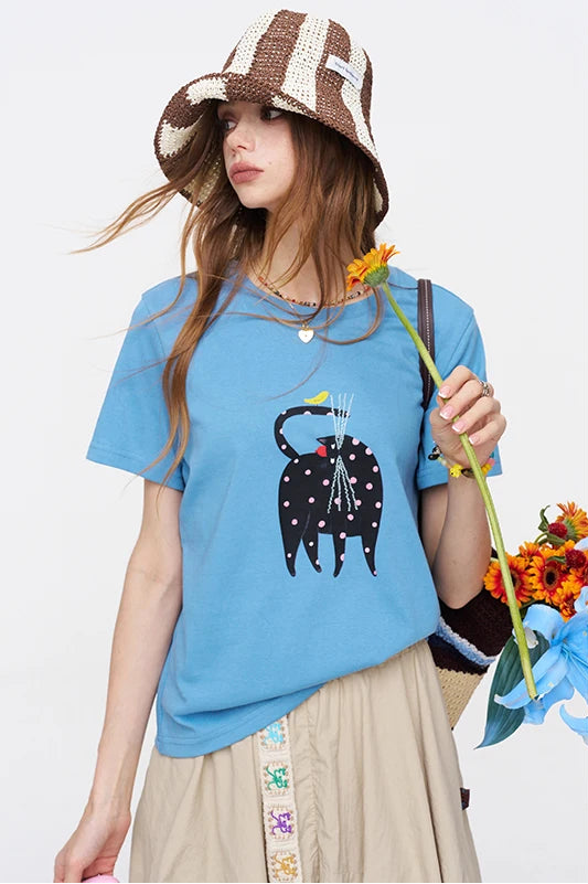 A woman wearing a light blue graphic t-shirt with a black cat design, a brown and beige striped bucket hat, and a beige skirt, holding a bouquet of orange flowers against a plain white background.