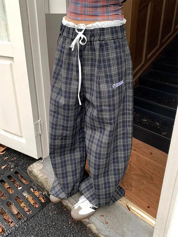 A person wearing very loose, wide-leg plaid lounge pants in navy, gray, and white, standing at an open doorway.