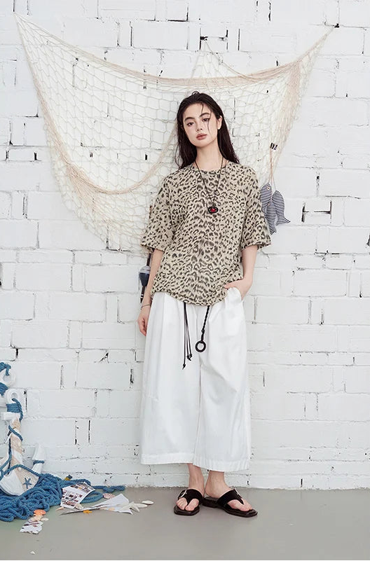 A woman stands against a white brick wall wearing a beige and black leopard-print short-sleeve blouse with puff sleeves, paired with white wide-leg pants and black sandals. A cream fishing-net textile hangs behind her, with a few loose fabric pieces on the floor at her side.