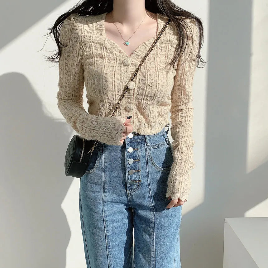 A woman wearing a beige cable-knit long-sleeve button-front cardigan and high-waisted blue jeans, with a small black crossbody bag, photographed against a sunlit white wall.