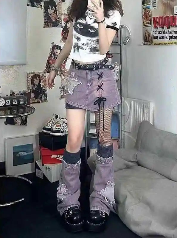 A person standing in a room wearing a white graphic T-shirt, a distressed lavender denim mini skirt with lace-up details, fishnet stockings, gray socks, and black platform shoes.