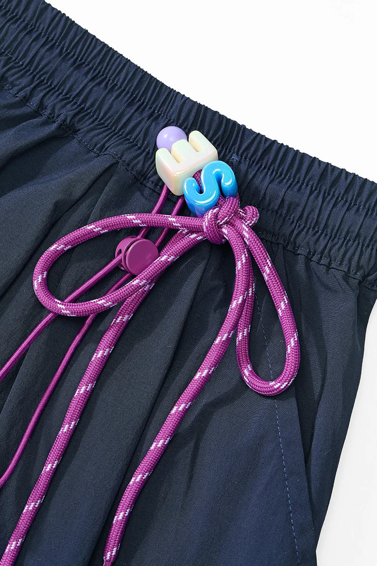 Close-up of dark navy shorts with a purple drawstring and colorful plastic toggle beads on a white background.