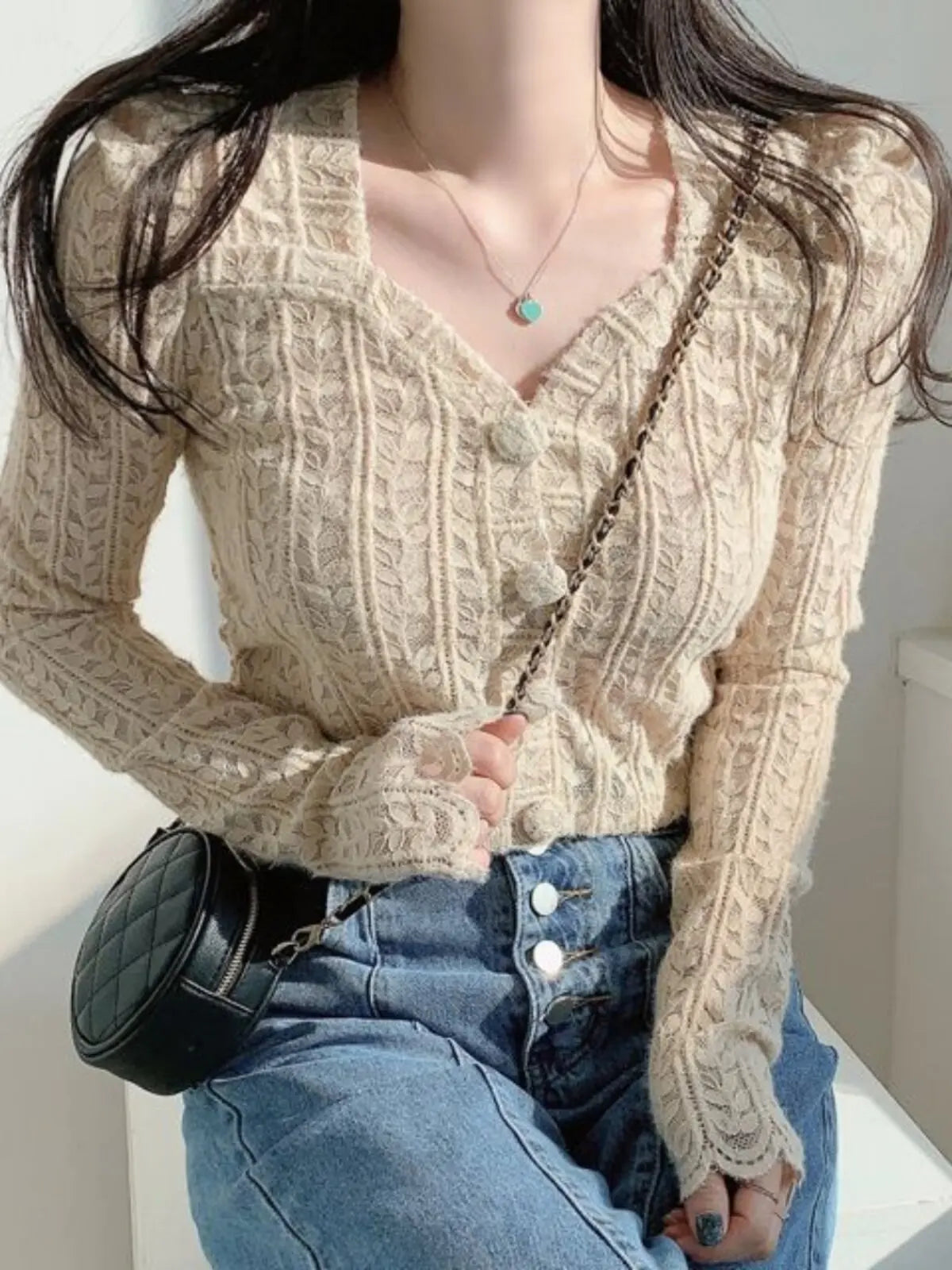 A woman wearing a cream-colored, long-sleeve cable-knit cardigan with a V-neck and decorative buttons, paired with blue jeans and a small black quilted crossbody bag.