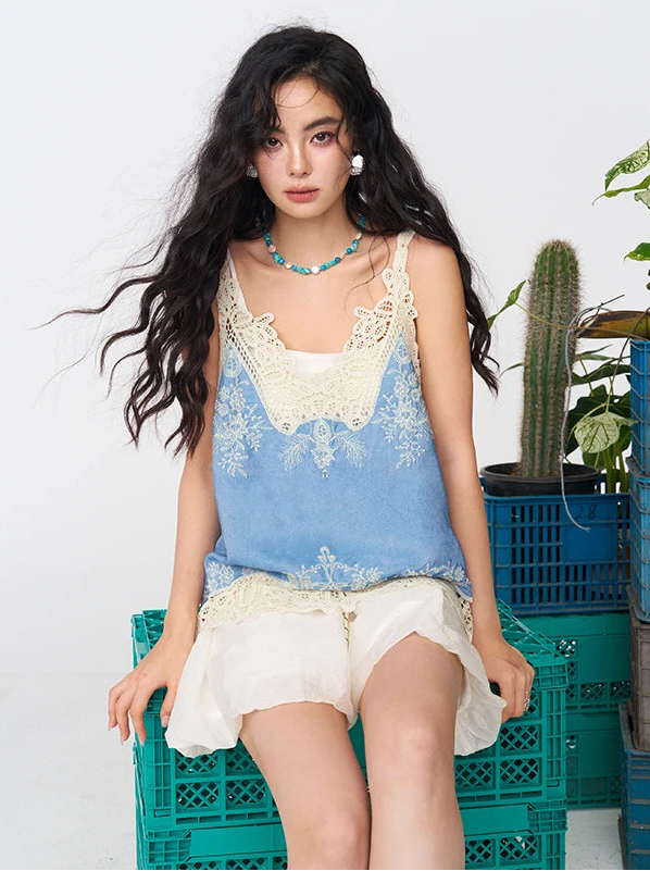 A woman models a light blue sleeveless top with cream lace trim and matching cream lace shorts, sitting on a teal crate in a studio setting with plants in the background.
