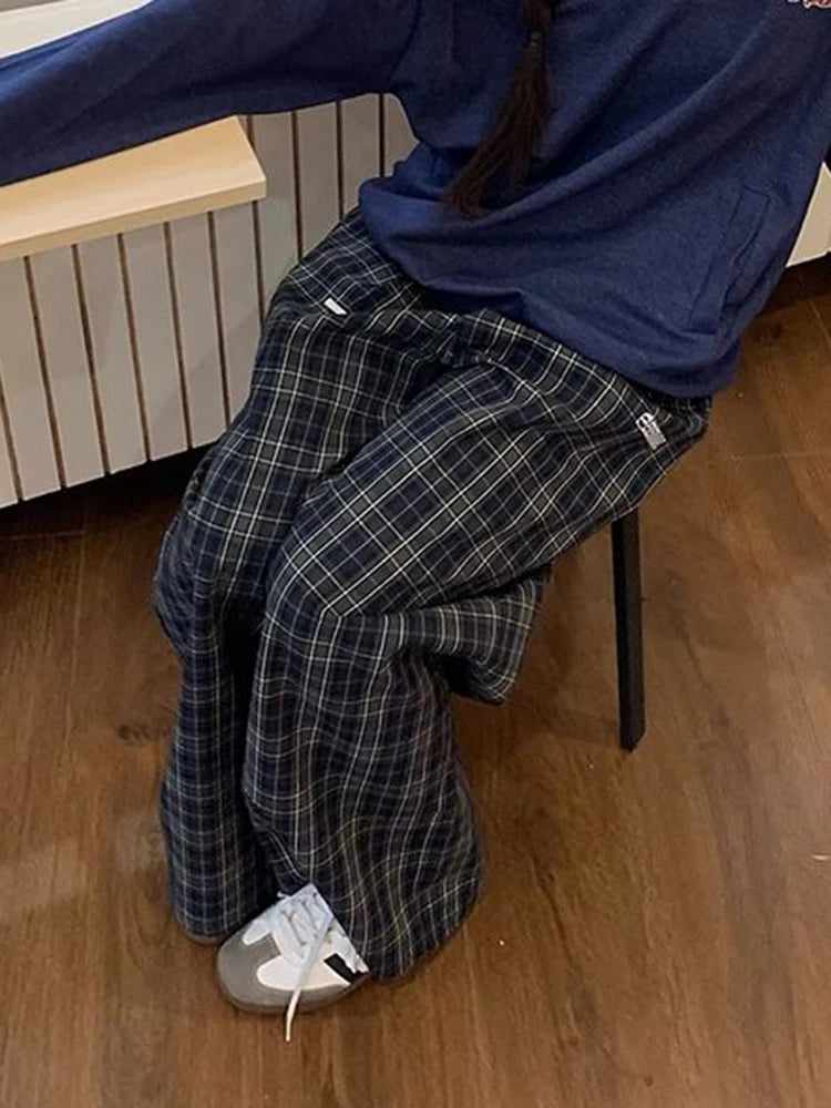 A person seated on a chair wearing loose dark plaid pants with a navy top and white sneakers in an indoor room with wood flooring.