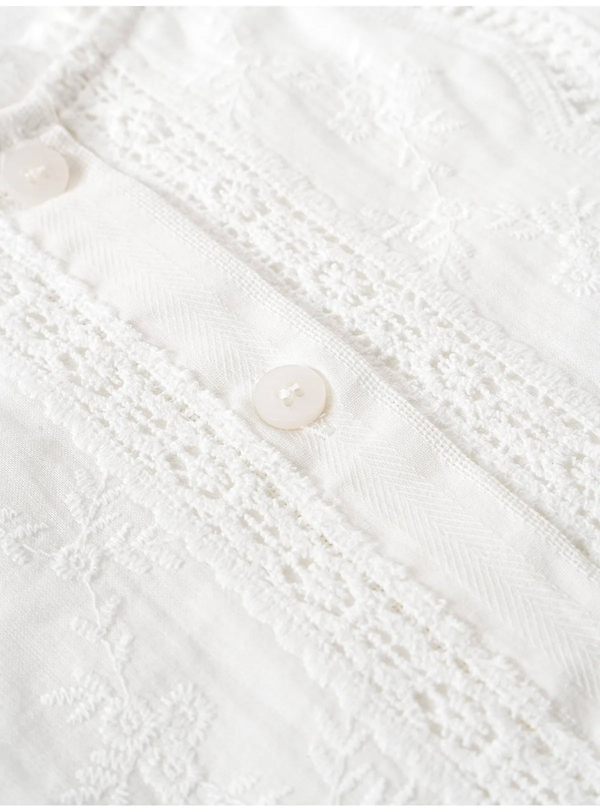 Close-up of a white embroidered fabric with a lace panel and small white buttons.