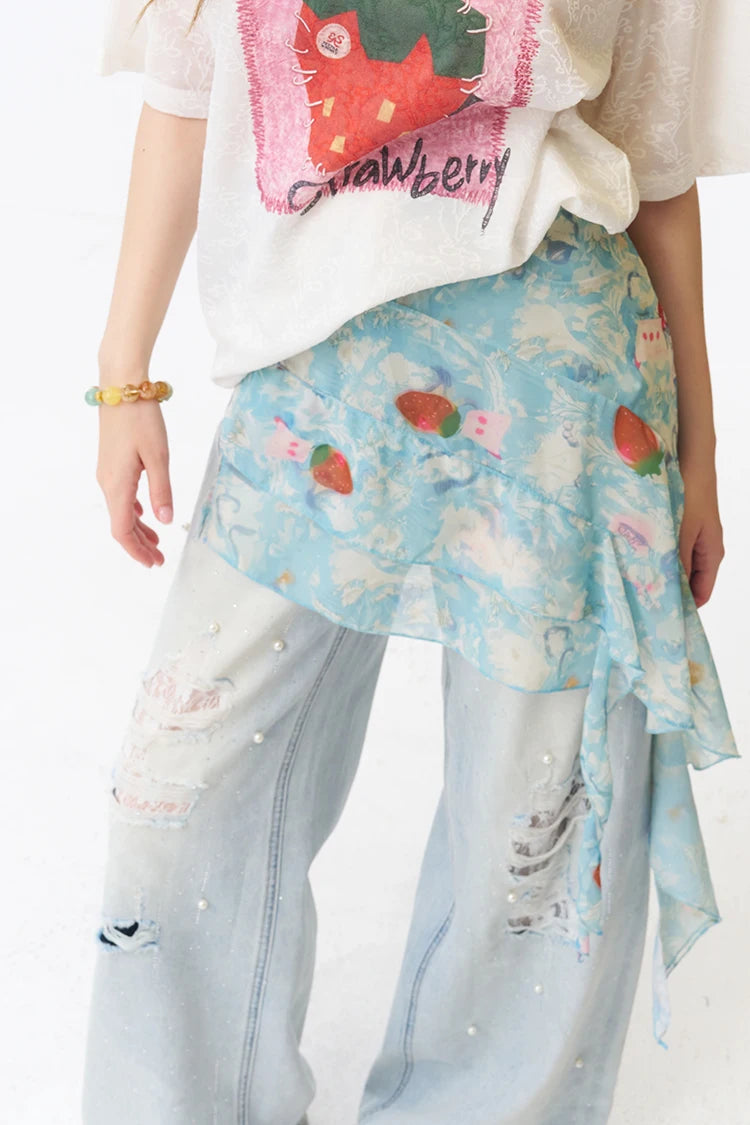 A person wearing a light blue floral sarong-style wrap with strawberry motifs over light-wash distressed jeans and a white graphic T-shirt, photographed against a plain white background.