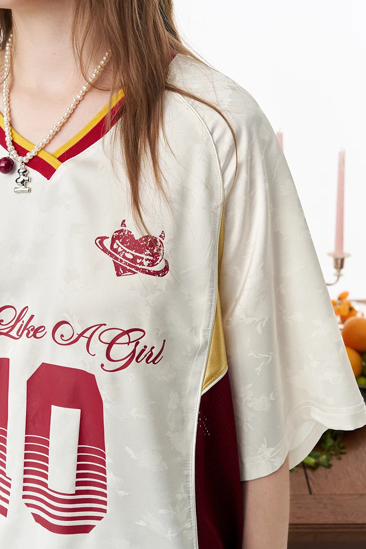 A close-up of a model wearing a cream-colored short-sleeve jersey with red and yellow accents, featuring a heart-and-rainbow graphic and the text 'Like A Girl' and '10'.
