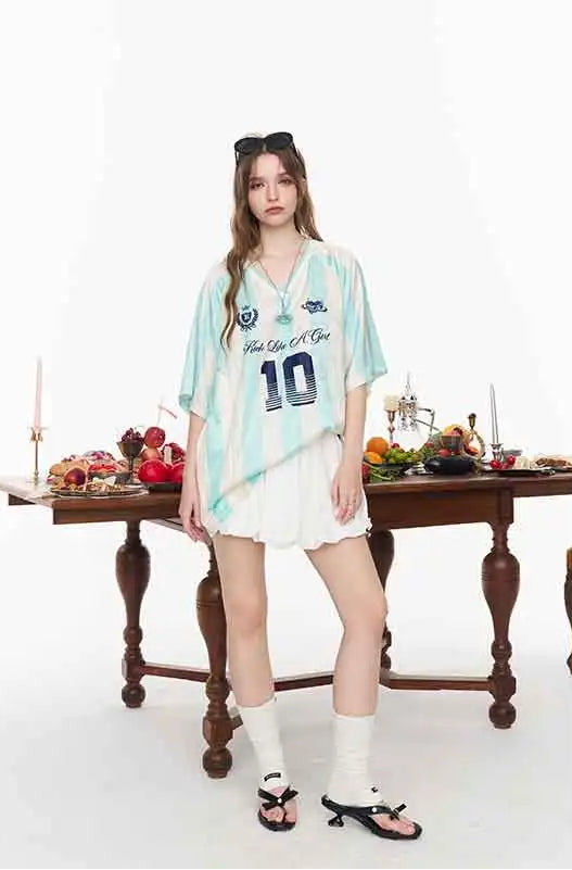A model stands in a studio setting wearing a light blue and white vertically striped oversized short-sleeve jersey dress with 'The Loli Club' and the number '10' printed on the front, paired with white shorts, white socks, and black sandals. A dark wooden table with decorative items is positioned behind her against a plain white background.