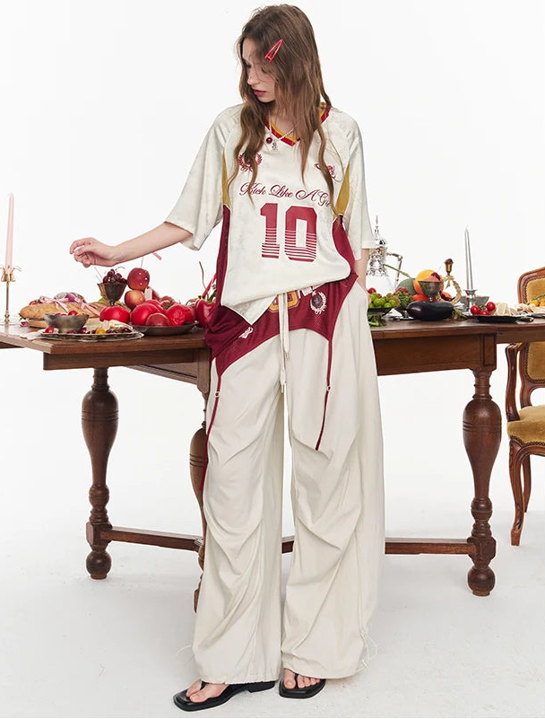 A woman models a cream and maroon two-piece lounge set with a graphic tee and matching wide-leg pants, standing in front of a wooden dining table decorated with fruit and candles.