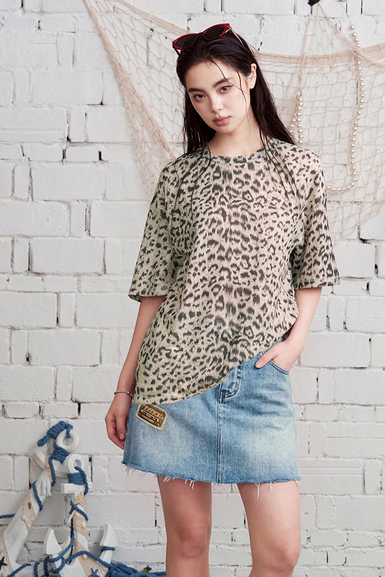A woman modeling a short-sleeve leopard print top with a light beige base and dark brown spots, paired with a denim mini skirt, standing against a white brick wall with nautical rope decorations.