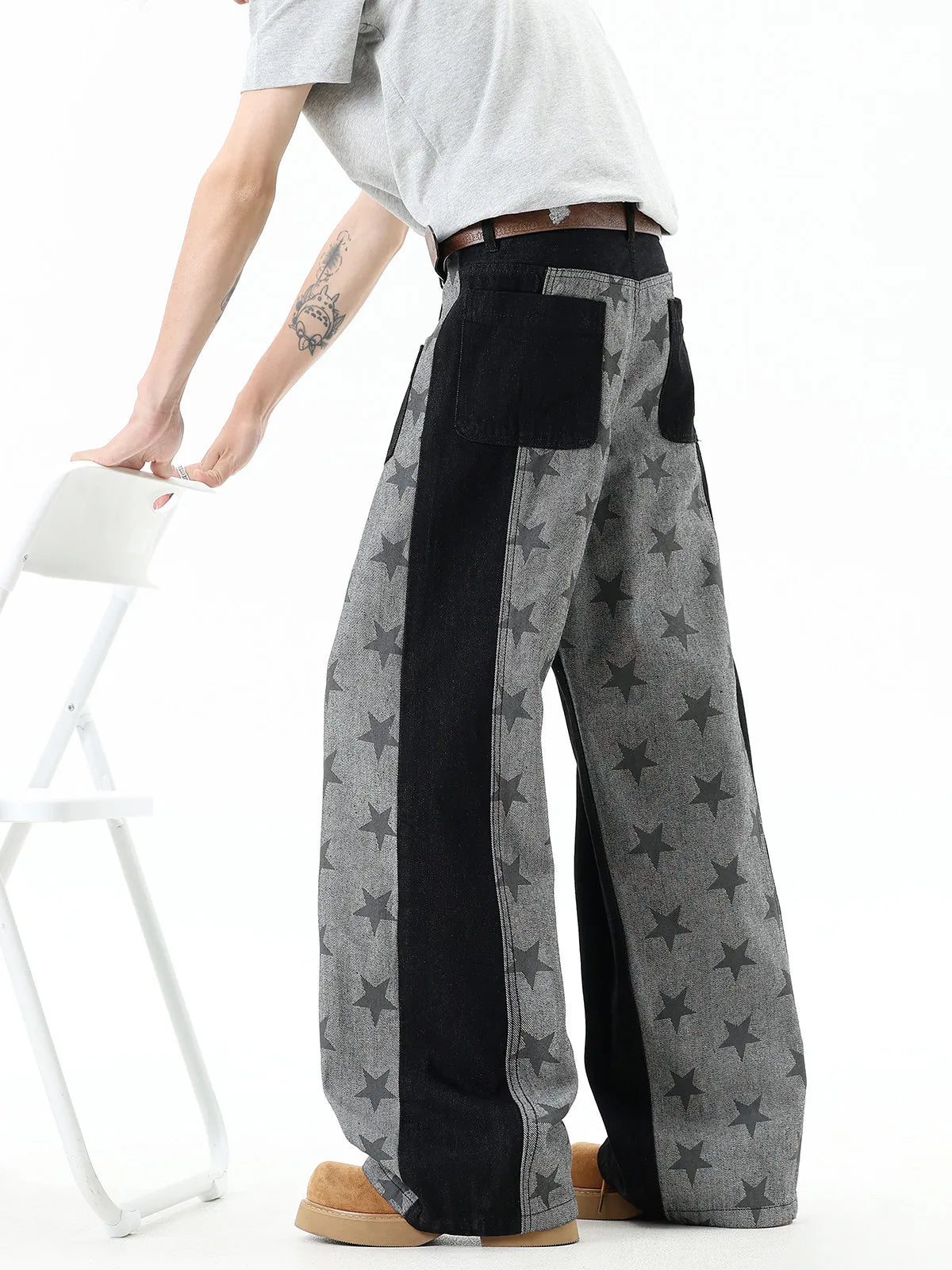 A person wearing wide-leg gray pants with black star prints and solid black side panels, paired with a light gray t-shirt and tan boots, standing next to a white folding chair.