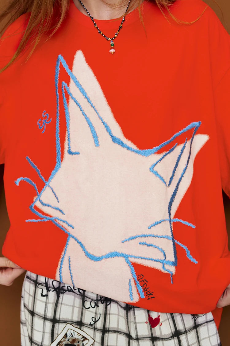 A close-up of a person wearing a bright orange-red crewneck sweatshirt with a large white cat face graphic outlined in blue. The person is also wearing a black and white plaid skirt and a silver necklace.
