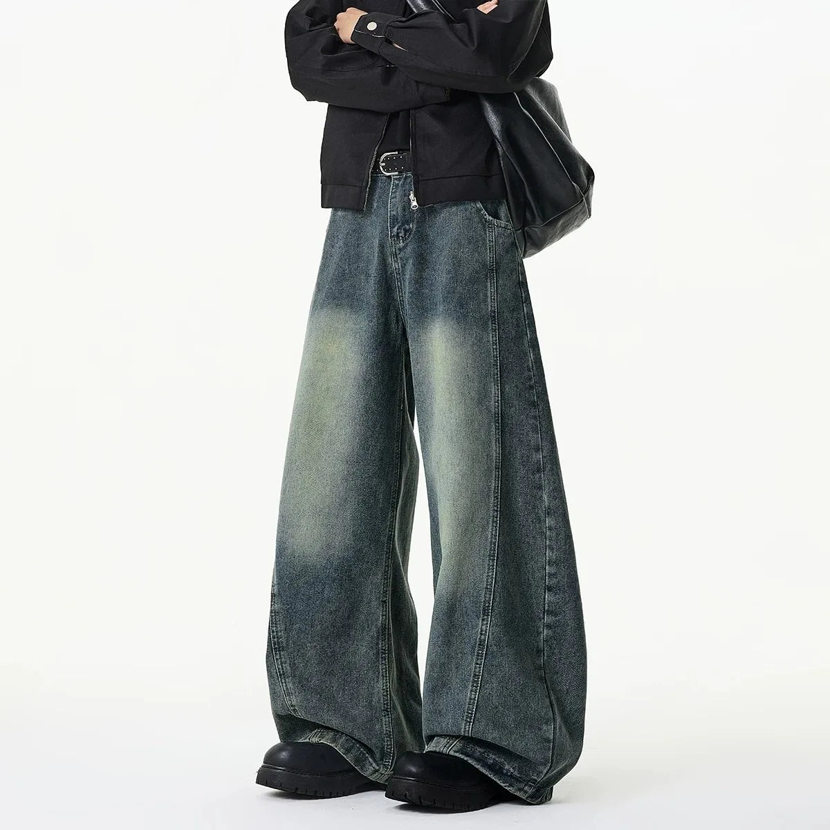 A person wearing wide-leg faded blue denim jeans, a black jacket, and black shoes, standing against a plain white studio background.