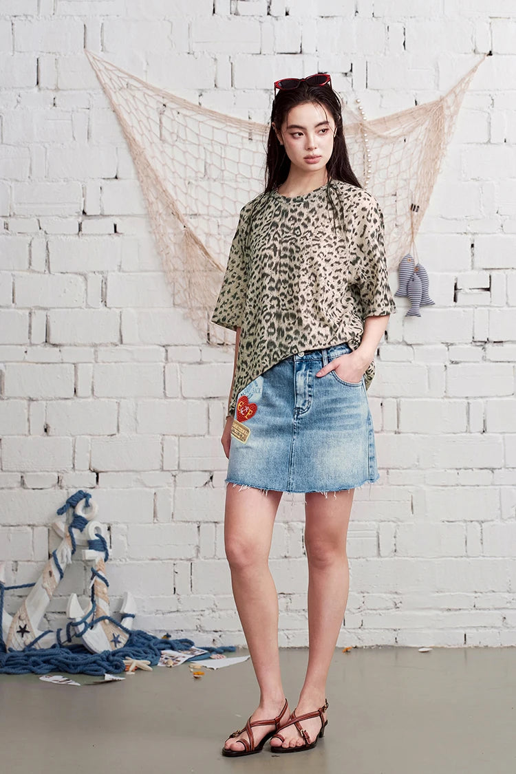 A woman stands against a white brick wall wearing a beige leopard-print short-sleeve top and a light-wash denim mini skirt with colorful patchwork details on the front. She is wearing brown strappy sandals, and a fishing net and blue fabric decor hang on the wall behind her.