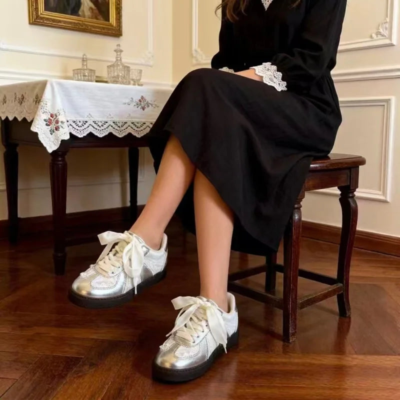 A person seated on a wooden stool in an indoor room, wearing a long black dress with white lace trim at the cuffs and shiny metallic silver low-top sneakers with oversized white laces. The room has a dark wood floor, a table with a white lace-trimmed tablecloth, and cream paneled walls.