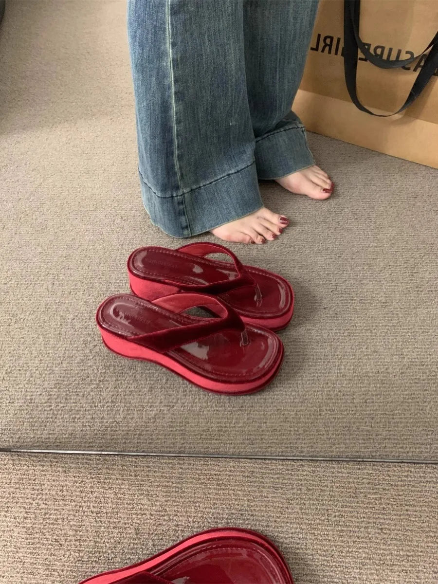 Red translucent flip-flop sandals placed on a beige carpet next to a person's bare feet wearing blue jeans, with a brown shopping bag in the background.