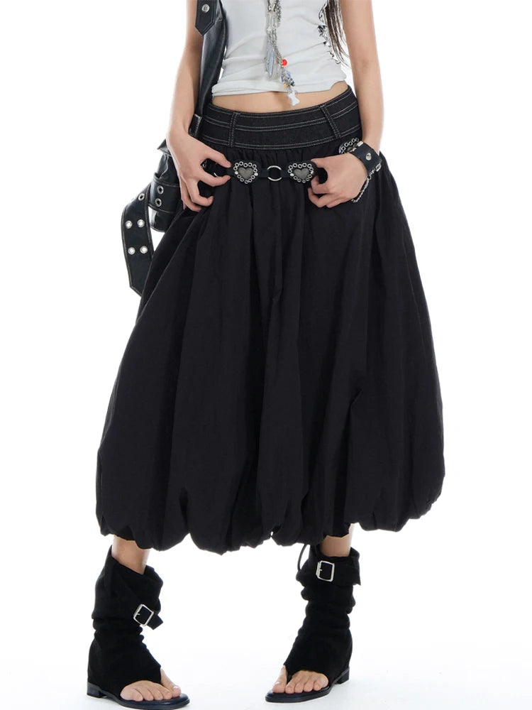 A person wearing a voluminous black harem-style skirt with a wide waistband, decorative belt hardware, a black studded shoulder bag, and black ankle boots against a plain white studio background.