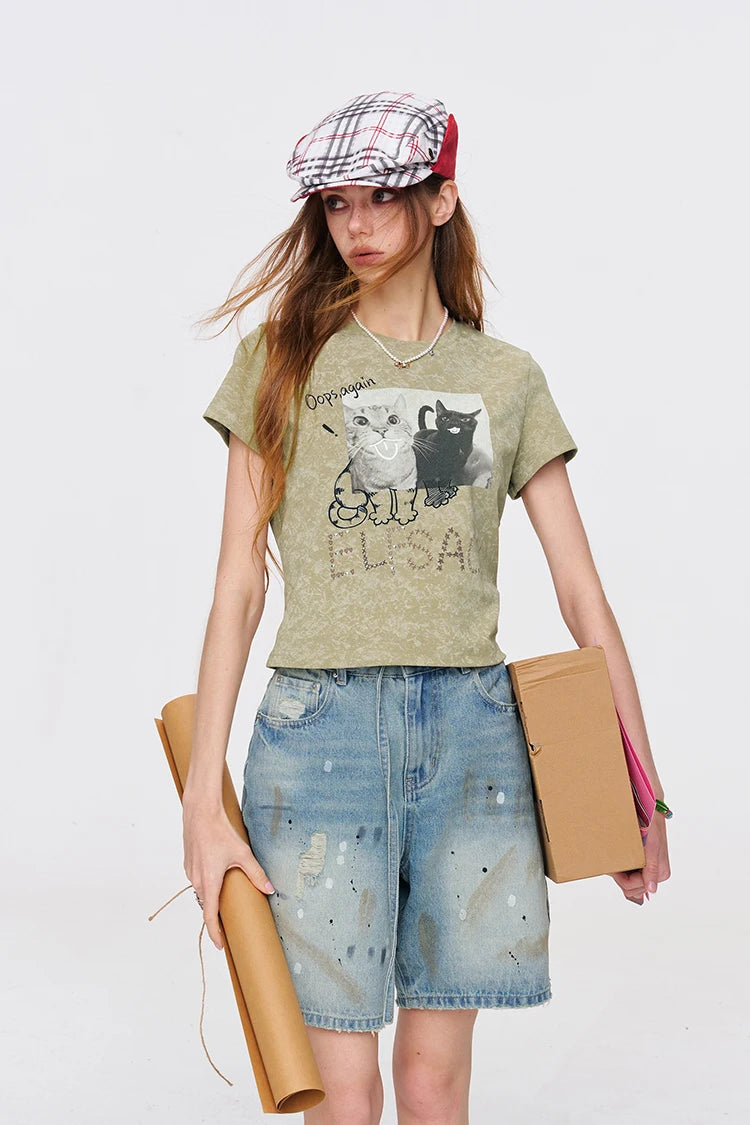 A model wearing a light olive green graphic t-shirt with a cat illustration, paired with distressed denim shorts, a plaid flat cap, and holding brown cardboard boxes against a plain white studio background.