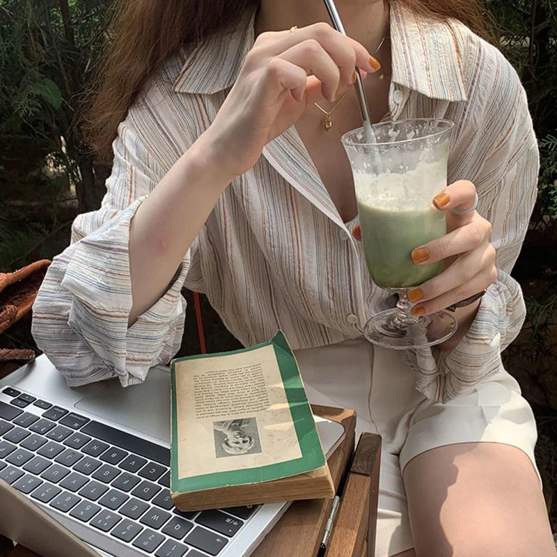 Person holding a green smoothie with a laptop and book on a table outdoors.
