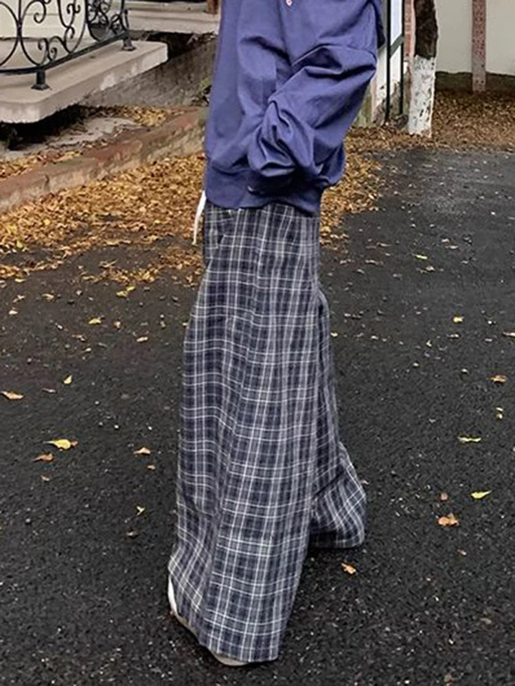 A person outdoors wearing a loose blue sweatshirt and very wide-leg plaid trousers in dark gray, white, and light blue, standing on a paved path with fallen leaves.