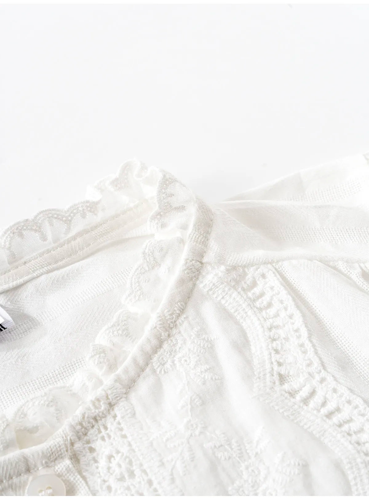 Close-up of a white lace and embroidered blouse with ruffled trim and a buttoned neckline on a white background.