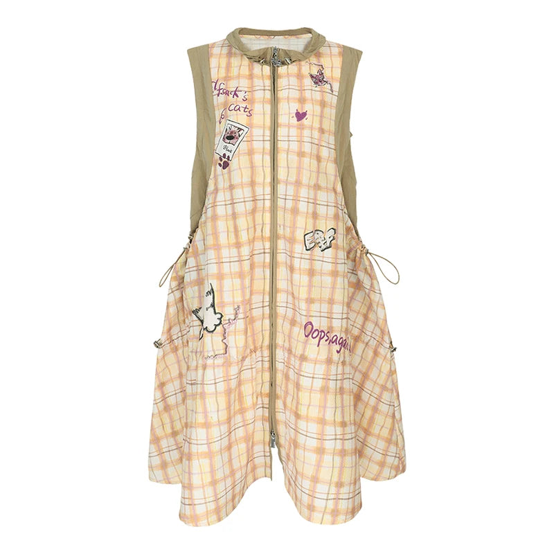 A sleeveless zip-front dress with a light peach and beige plaid pattern, decorated with various cartoon patches, embroidered text, and drawstring details, shown against a plain white background.