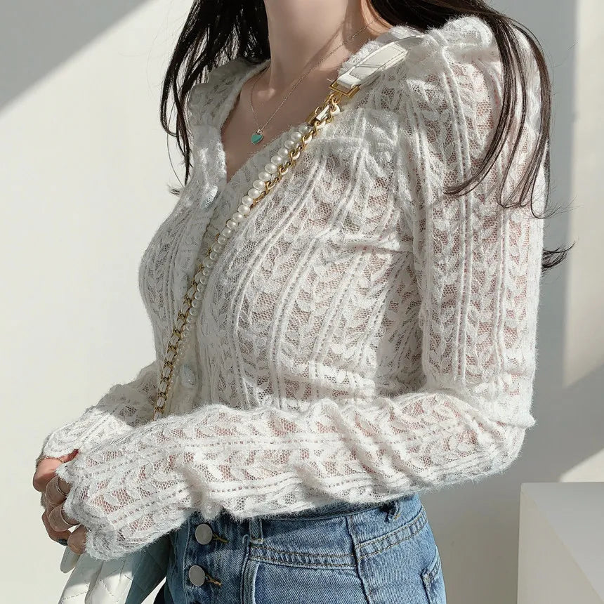 A woman wearing a white cable-knit long-sleeve sweater with a V-neckline, paired with blue jeans and a gold chain strap bag, set against a bright white wall with soft shadows.