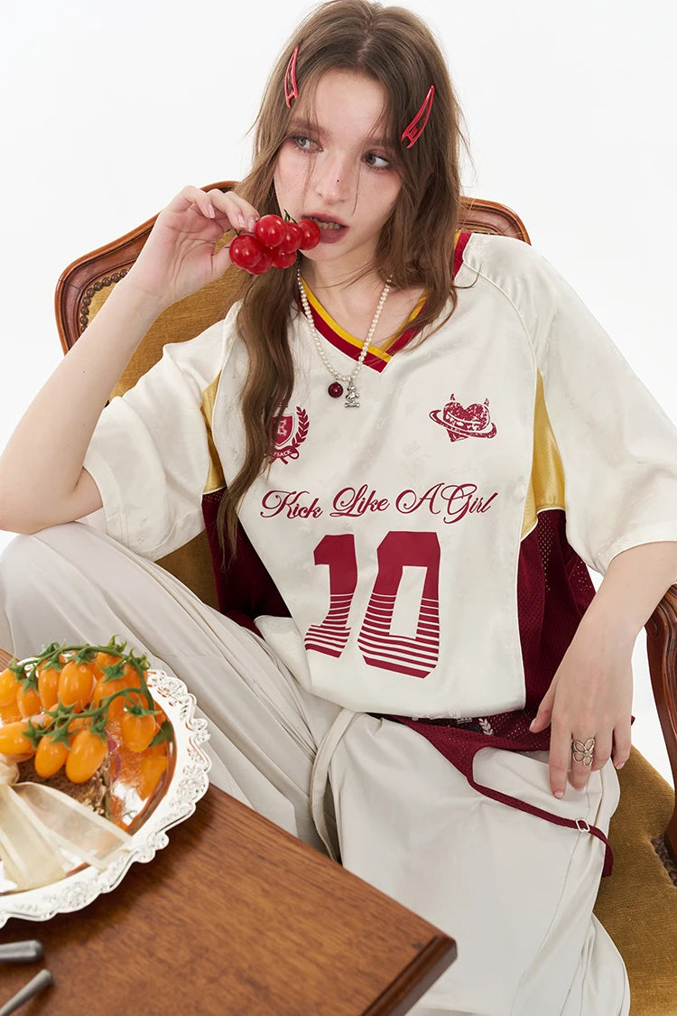 A woman sitting in a wooden chair wearing a cream-colored sports jersey with red and maroon accents, featuring the text 'F*ck Like A Girl' and the number '10' on the front. She is holding red berries near her mouth, with a tray of small orange fruits on a table in the foreground.