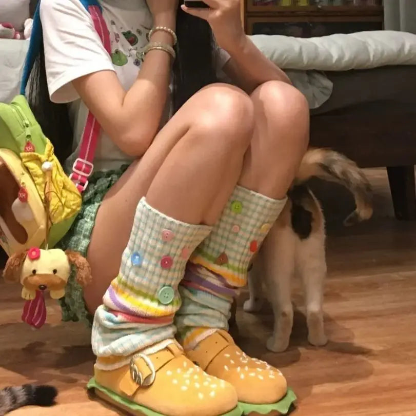 A person sitting on a wooden floor wearing pastel striped leg warmers with colorful buttons and yellow slip-on shoes, with a cat standing nearby in a home setting.