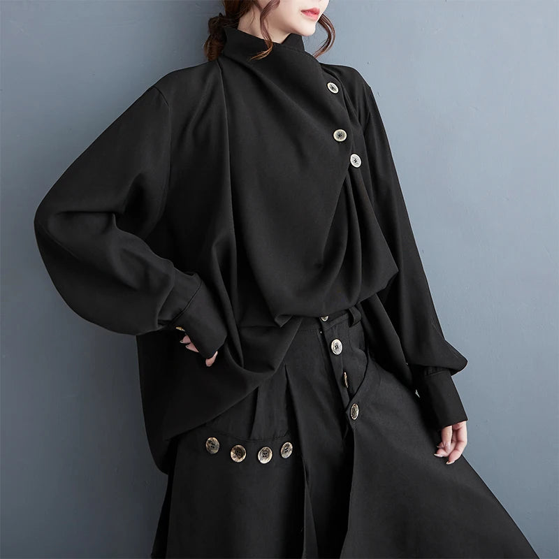A model wearing a black high-neck, long-sleeve blouse with asymmetrical decorative buttons and matching black trousers with button details, set against a plain gray wall.
