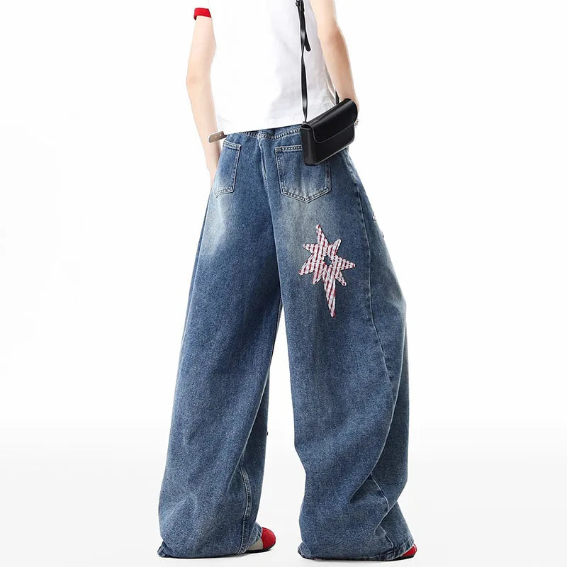 A model from the waist down wearing wide-leg blue denim jeans with a pink and white checkered star patch on the left leg, styled with a white sleeveless top and a small black crossbody bag against a white background.