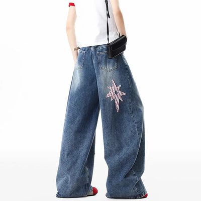 A model from the waist down wearing wide-leg blue denim jeans with a pink and white checkered star patch on the left leg, styled with a white sleeveless top and a small black crossbody bag against a white background.