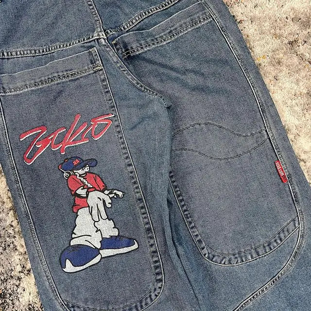 Close-up of the back pocket of blue denim jeans featuring a large embroidered cartoon character and red script text on the pocket, laid flat on a textured beige surface.