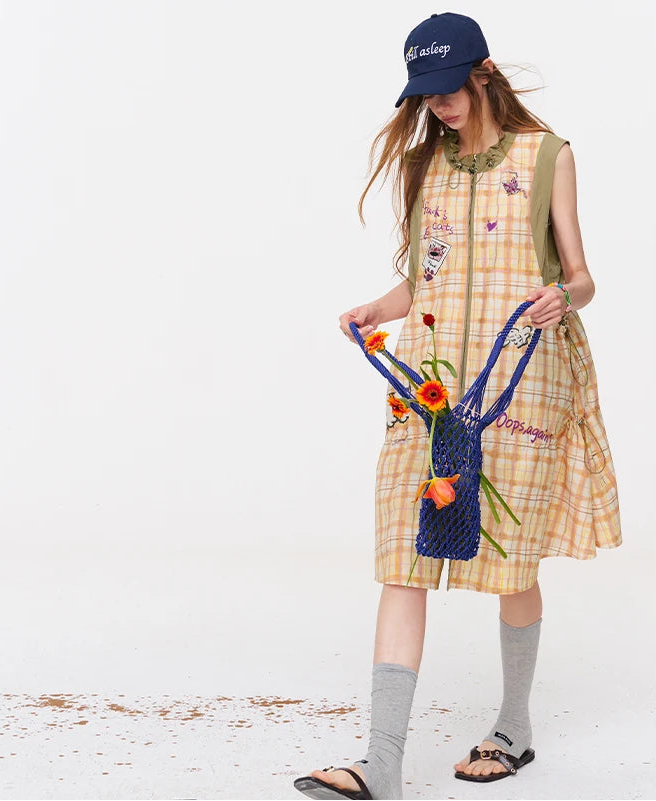 A woman models a sleeveless yellow and white plaid dress with colorful embroidered details, styled with a navy blue baseball cap, gray knee-high socks, sandals, and a blue flower bag against a plain white studio background.