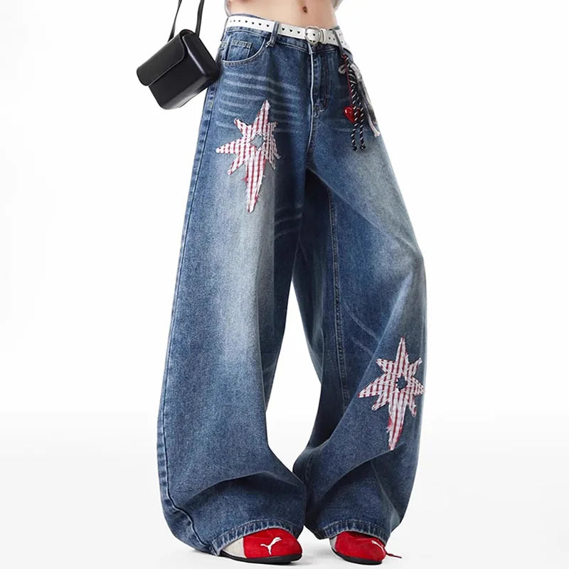 A person wearing very wide-leg blue denim jeans with large pink-and-white gingham star patches on the thighs and lower legs, styled with a white belt, a small black shoulder bag, and red shoes against a plain white background.