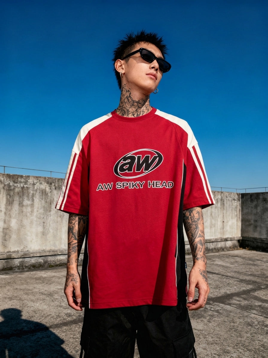 A tattooed man wearing a red and white AW Spiky Head t-shirt and black sunglasses, standing outdoors against a clear blue sky and concrete wall.